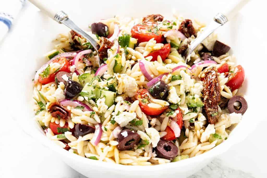 A bowl of Greek Orzo Pasta Salad with cherry tomatoes, red onion, cucumbers, Kalamata olives, sun-dried tomatoes, fresh herbs, and tangy feta cheese, served with two utensils.