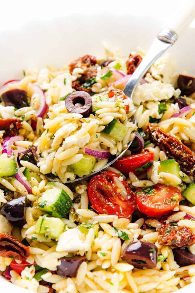 A close-up of Greek Orzo Pasta Salad with cherry tomatoes, cucumber, red onion, Kalamata olives, feta cheese, and herbs, with a fork lifting a portion from a white bowl.