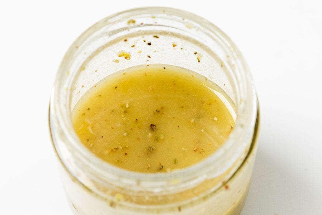 A close-up of an open glass jar filled with a light yellow, speckled liquid, likely a homemade vinaigrette for Greek Orzo Pasta Salad, against a white background.