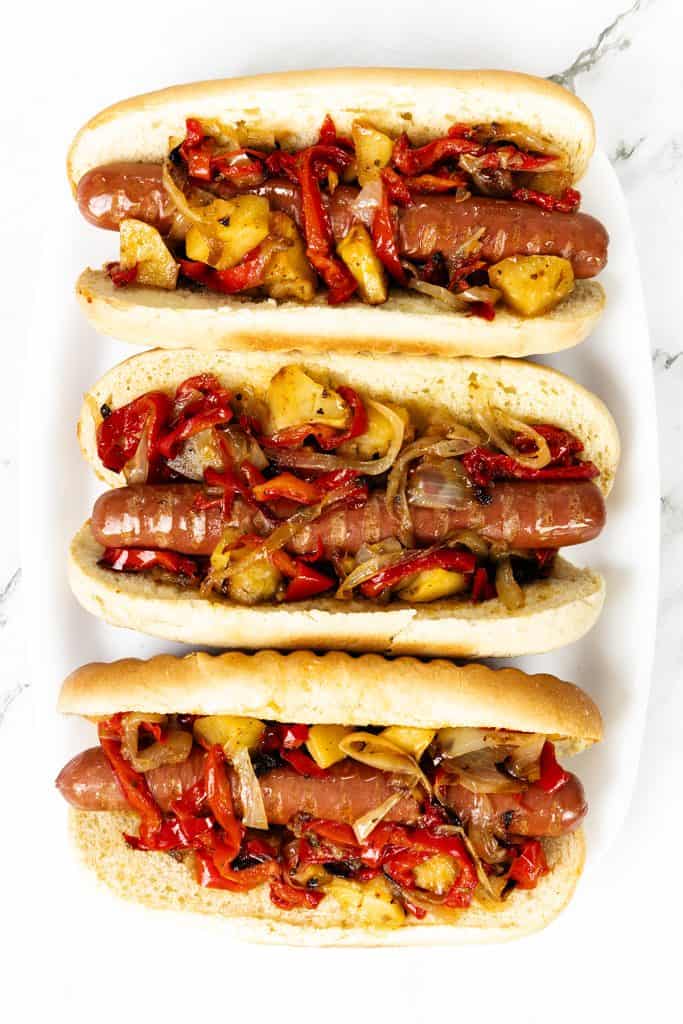 Three Italian Hot Dogs in buns, topped with sautéed bell peppers, onions, and pineapple slices, are arranged on a white plate against a white background.