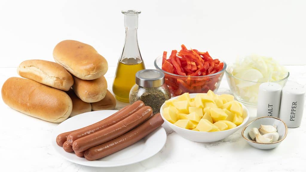 Hot dog buns, a plate with uncooked hot dogs, a bowl of cubed potatoes for Italian Hot Dogs, sliced red bell pepper, sliced onion, a bottle of oil, seasoning shaker, salt and pepper shakers, and garlic on a white surface.