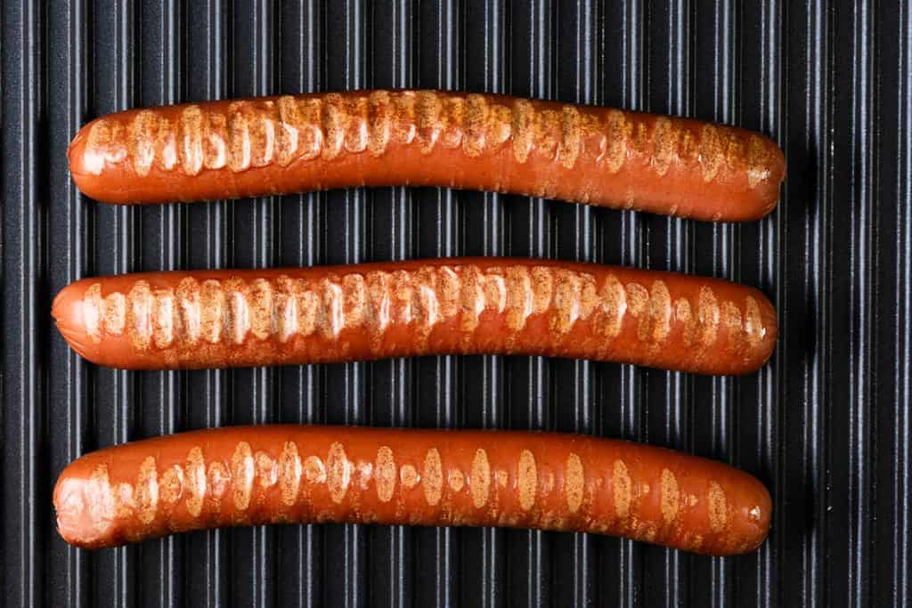 Three Italian Hot Dogs with seared marks are lined up horizontally on a black ridged grill surface, showcasing their irresistible grilled perfection.