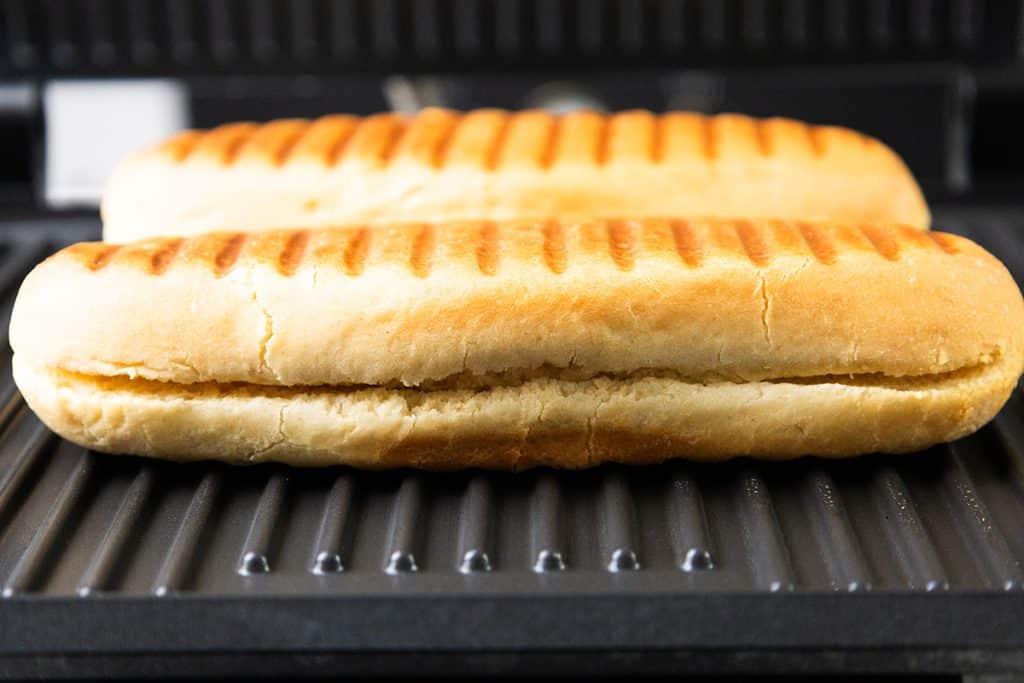 Two sandwich rolls with browned grill marks, filled to make classic Italian Hot Dogs, are placed side by side on a panini press, ready for toasting.