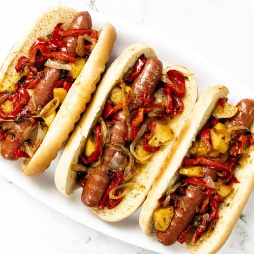 Three Italian Hot Dogs in buns are topped with sautéed onions and red bell peppers, arranged on a white plate. French fries are partially visible on the sides of the image.