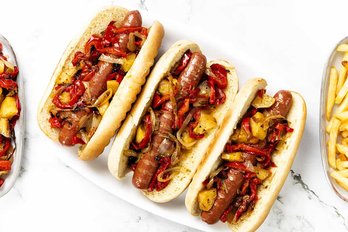 Three Italian Hot Dogs in buns are topped with sautéed onions and red bell peppers, arranged on a white plate. French fries are partially visible on the sides of the image.
