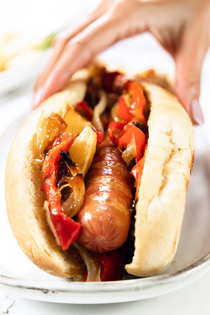 A close-up of an Italian Hot Dog in a bun, topped with sautéed onions and red bell peppers. A hand is holding the side of the bun on a white plate.