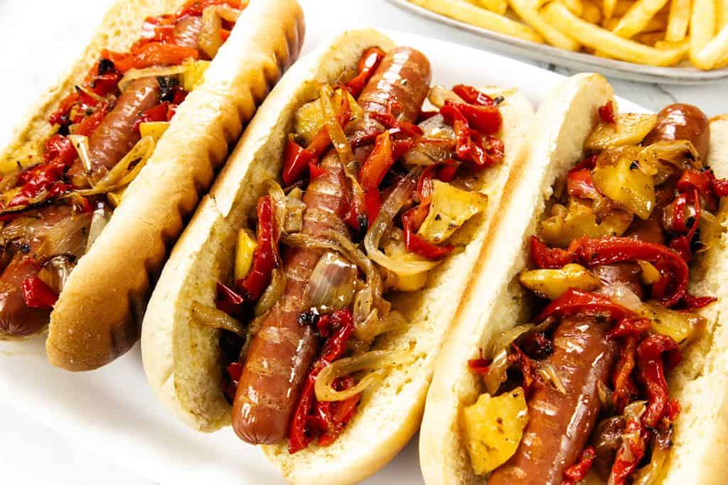 Three Italian Hot Dogs in buns are topped with sautéed onions and red and yellow bell peppers. A portion of golden French fries is visible in the background.