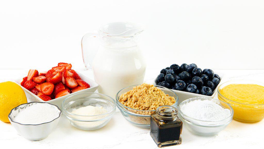A variety of ingredients for a Lemon Dessert are displayed on a white surface, including cut strawberries, blueberries, a lemon, granulated sugar, almond meal, powdered sugar, vanilla extract, milk in a jug, and a bowl of yellow liquid.