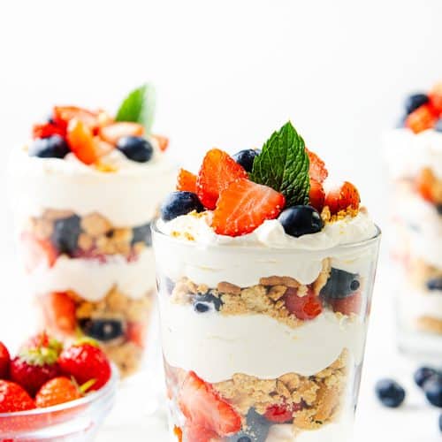 Three layered lemon cream parfaits in clear glasses, filled with whipped cream, crumbled cookies, strawberries, and blueberries, and topped with mint leaves. A bowl of fresh strawberries and scattered blueberries are nearby for this refreshing parfait recipe.