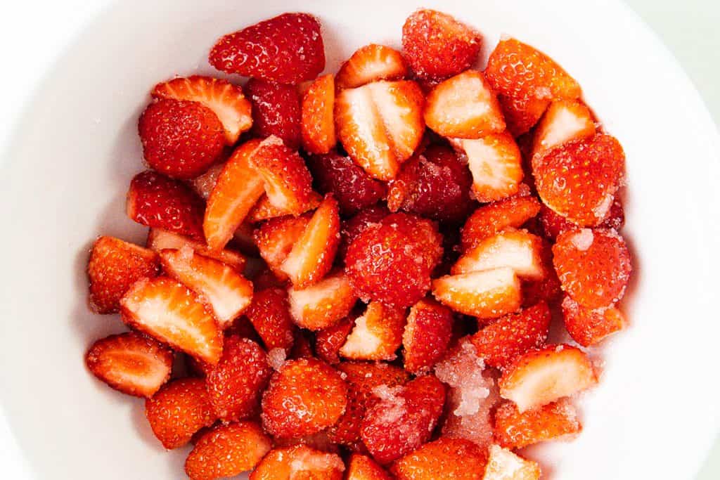 A white bowl filled with halved and sliced fresh strawberries, some sprinkled with sugar&mdash;perfect for topping parfaits or enjoying as a simple lemon dessert.