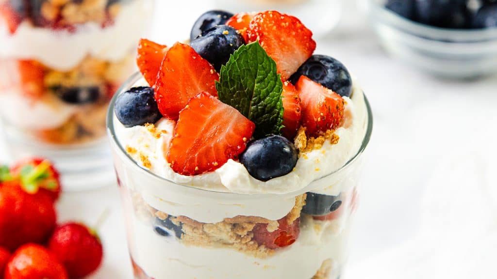 A glass filled with layers of whipped cream, crumbled cookies, strawberries, and blueberries, topped with fresh strawberry slices, whole blueberries, and a mint leaf&mdash;a vibrant parfait recipe. Other berries and parfaits are visible in the background.