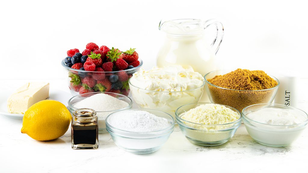 A variety of baking ingredients on a white surface, perfect for an easy dessert like a No-Bake Cheesecake—featuring fresh berries, butter, lemon, vanilla extract, milk, sugars, salt, flour, grated cheese, and whipped cream in glass bowls.