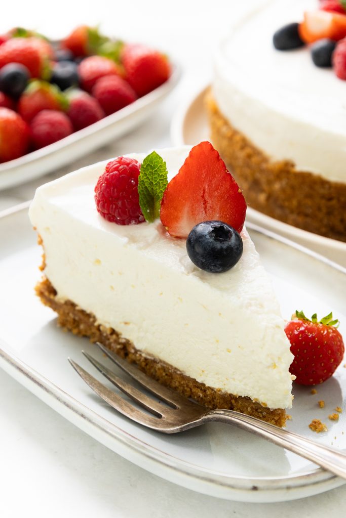 A slice of no-bake cheesecake with a graham cracker crust, topped with raspberry, blueberry, strawberry slice, and mint leaf, sits on a white plate with a fork. The rest of this easy dessert and mixed berries are in the background.