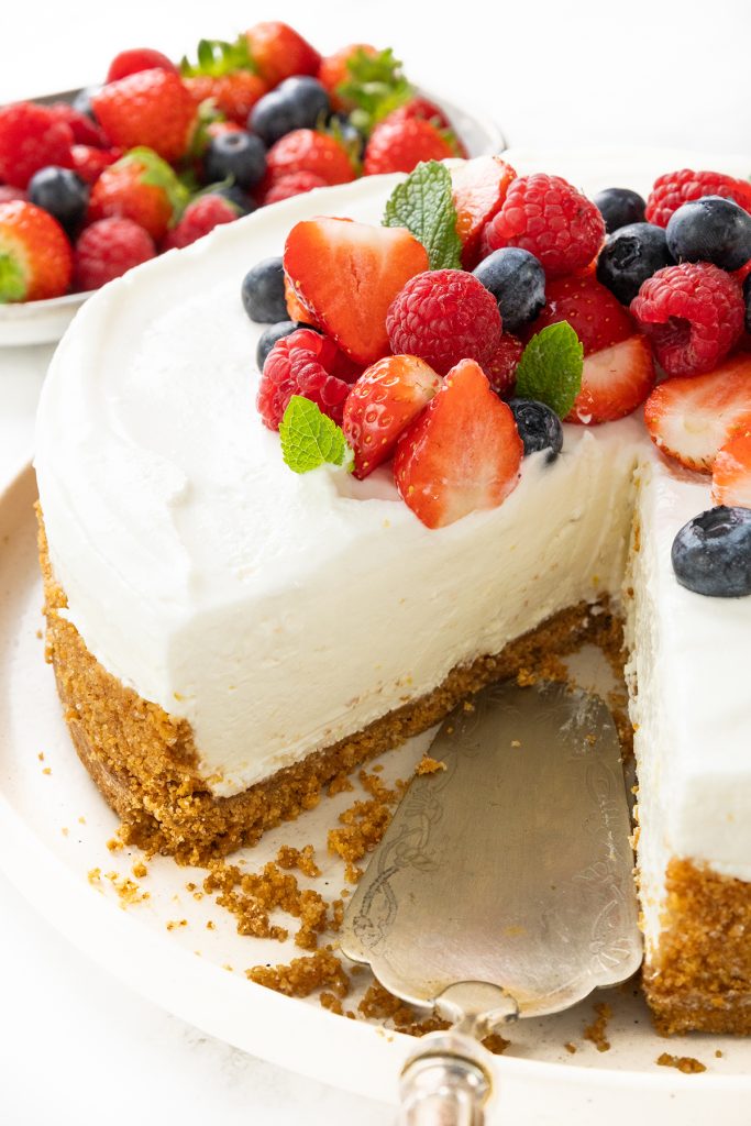 A creamy cheesecake with a graham cracker crust, topped with fresh strawberries, raspberries, blueberries, and mint leaves. This easy dessert has a slice served on a pie server, with a bowl of berries in the background.