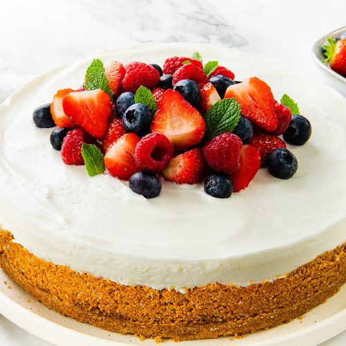 A classic cheesecake with a golden crust topped with whipped cream, fresh strawberries, blueberries, raspberries, and mint leaves—an easy dessert and perfect no-bake cheesecake recipe on a white plate surrounded by more berries.