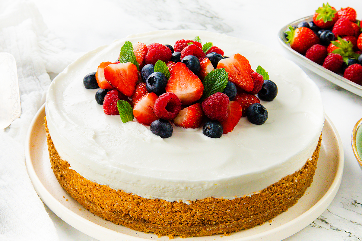 A classic cheesecake with a golden crust topped with whipped cream, fresh strawberries, blueberries, raspberries, and mint leaves—an easy dessert and perfect no-bake cheesecake recipe on a white plate surrounded by more berries.