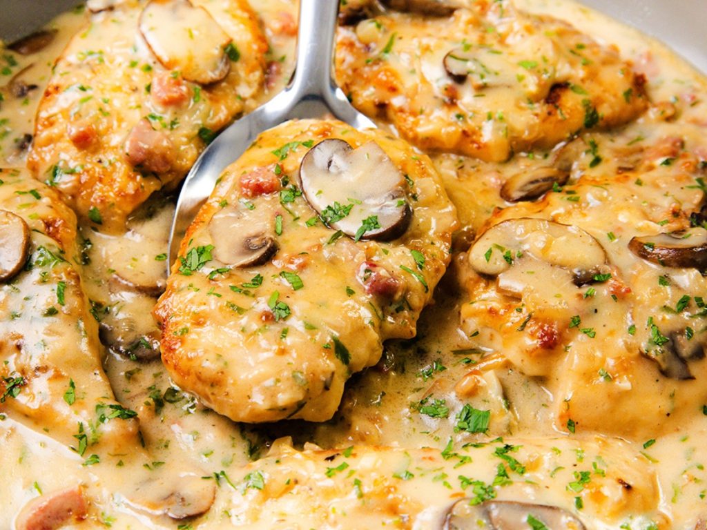 Golden-brown chicken breasts in a creamy mushroom sauce with bacon bits, garnished with chopped parsley, with a spoon lifting one piece from the pan. Sign up for our Free Recipe Newsletter to get delicious recipes like this delivered to your inbox!.