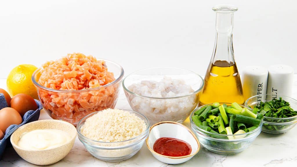 A selection of fresh ingredients on a white surface for Shrimp and Salmon Cakes, including diced salmon, shrimp, green onions, parsley, eggs, lemon, mayonnaise for spicy mayo, panko breadcrumbs, sriracha sauce, olive oil, salt, and pepper.