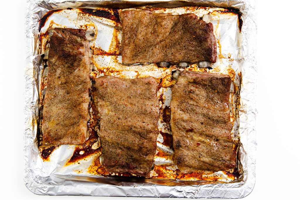 Four seasoned and cooked Asian Style Pork Ribs are laid out on a foil-lined baking sheet, with juices and spices visible on the foil.