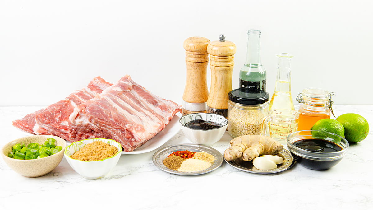 The ingredients for Asian style pork ribs arranged on a counter, including ribs, spices, sauces, lime, sesame seeds, and green onions.