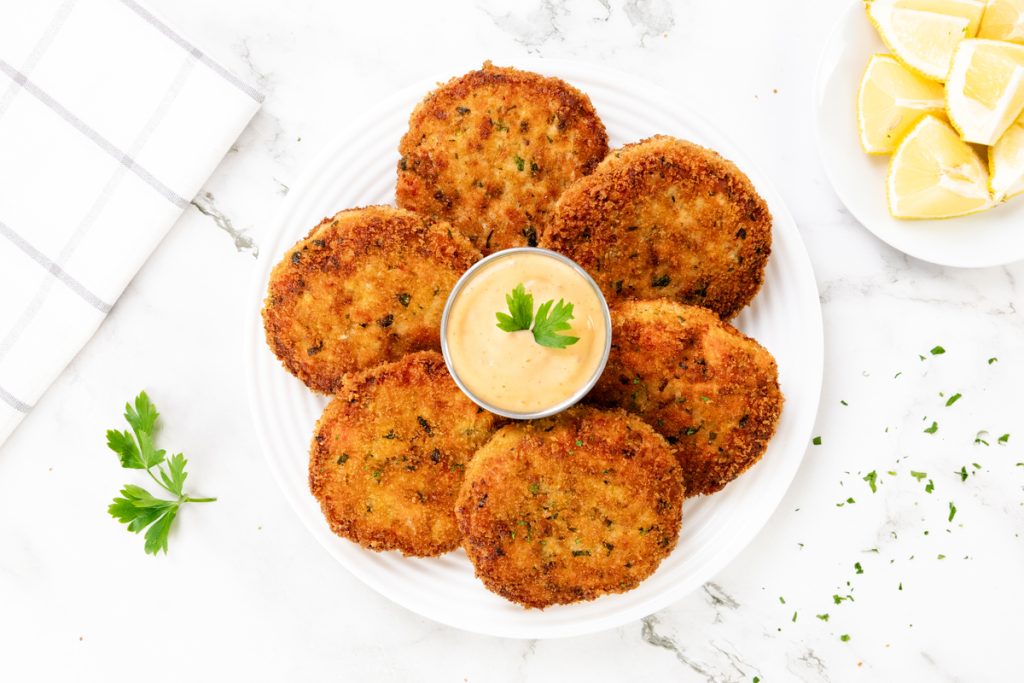 A white plate with six golden brown crab cakes arranged around a small bowl of spicy mayo, garnished with a parsley leaf. Lemon wedges and fresh parsley are on the side, atop a white marble surface.