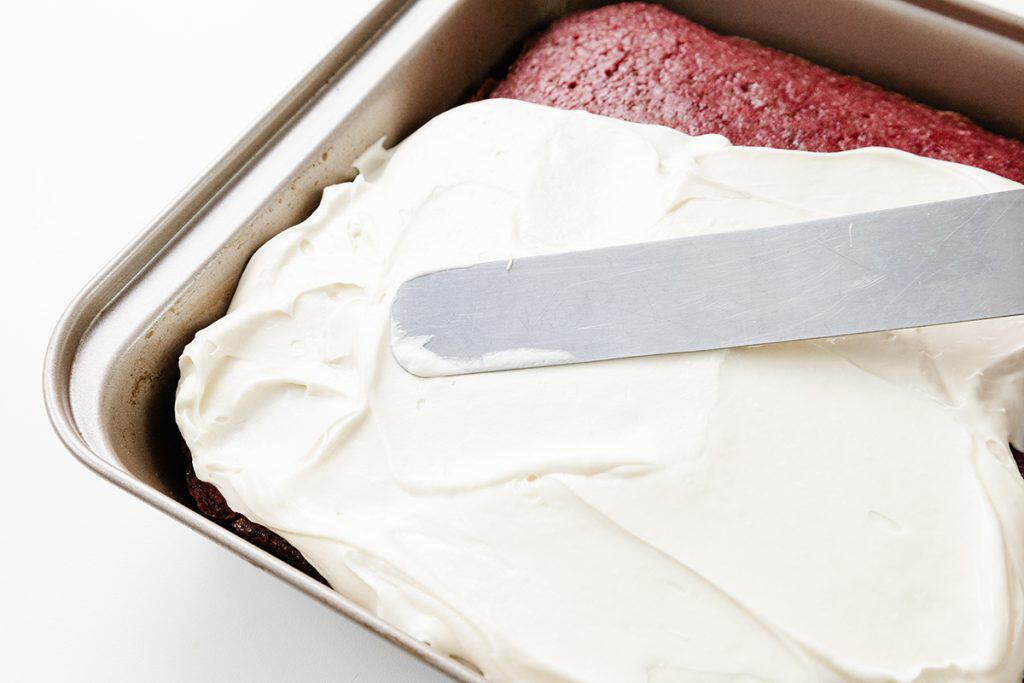 A metal spatula spreads creamy white frosting over a Red Velvet Sheet Cake in a rectangular baking pan.