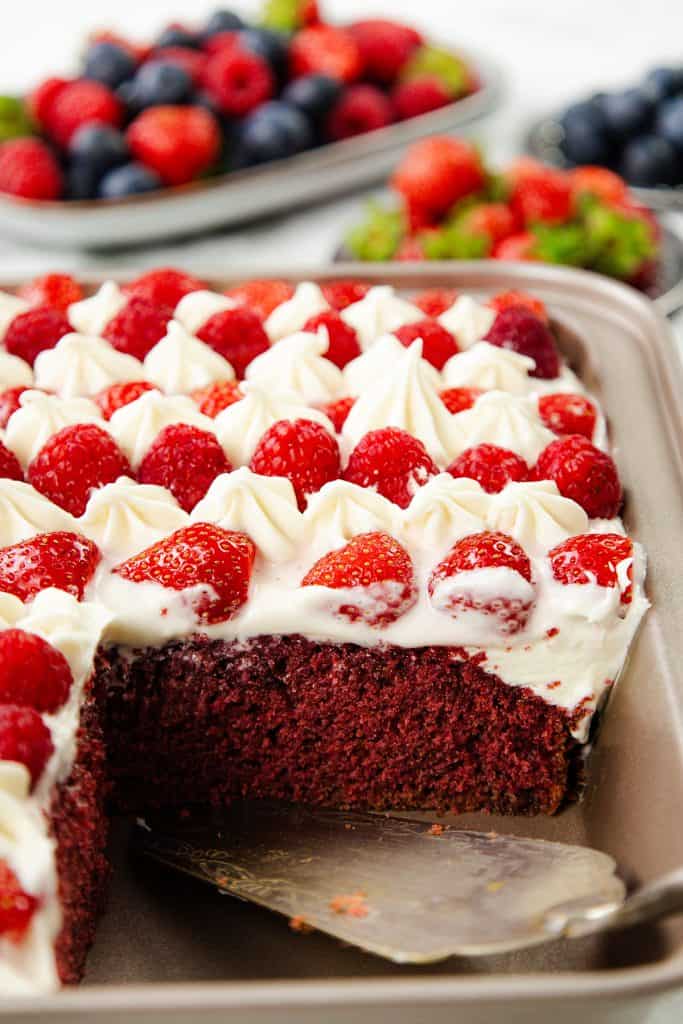 A rectangular Red Velvet Sheet Cake topped with white frosting and fresh strawberries, with a slice removed. Blurred plates of assorted berries are visible in the background, making this a perfect patriotic dessert for any celebration.