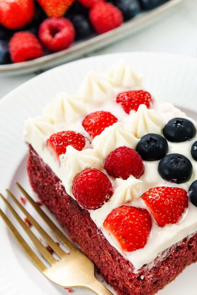 A slice of Red Velvet Sheet Cake topped with cream cheese frosting, fresh strawberries, raspberries, and blueberries sits on a white plate with a gold fork&mdash;an inviting patriotic dessert perfect for any celebration.