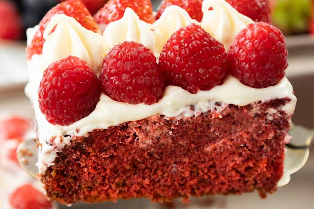 A close-up of a Patriotic Dessert, this Red Velvet Sheet Cake slice is topped with creamy white frosting and fresh raspberries. The moist cake boasts a vibrant red color and is being lifted on a spatula.