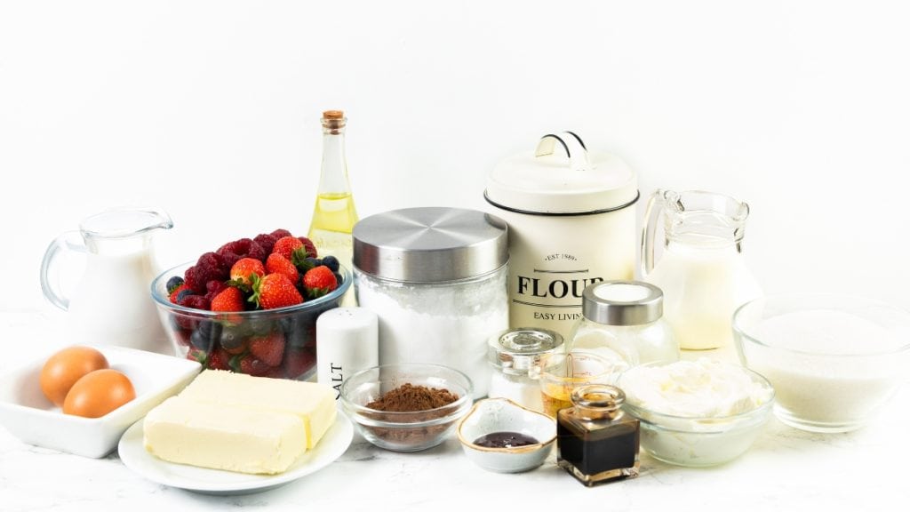 A variety of baking ingredients arranged on a white surface&mdash;perfect for creating a Red Velvet Sheet Cake&mdash;with eggs, butter, flour, sugar, milk, cream, cocoa powder, vanilla, salt, strawberries, and a bottle of oil.