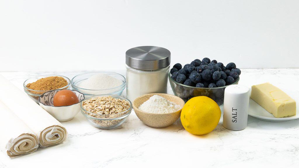 A marble countertop is set for Blueberry Pie: rolled pie crusts, an egg, brown sugar, white sugar, oats, flour, a lemon, salt, butter, a jar with a metal lid, and a bowl of fresh blueberries ready for baking.
