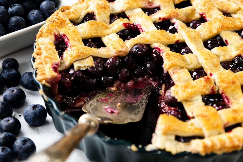 A close-up of a homemade Blueberry Pie with a golden lattice crust, a slice missing to reveal juicy blueberries inside. Fresh blueberries are scattered around the pie on a white surface.