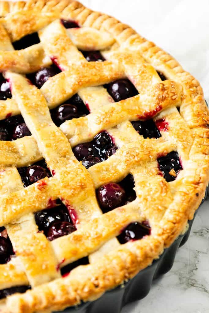 A close-up of a freshly baked cherry pie with a golden, flaky lattice crust, showing glossy cherries peeking through the gaps—reminiscent of the classic look of a homemade blueberry pie.