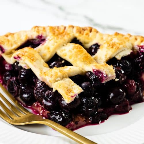A close-up of blueberry pie with a golden lattice crust, served on a white plate with a gold fork, against a marble background.