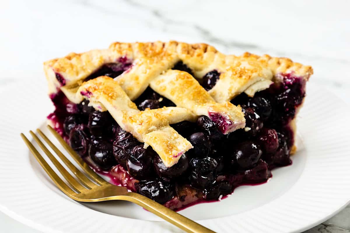 A close-up of blueberry pie with a golden lattice crust, served on a white plate with a gold fork, against a marble background.