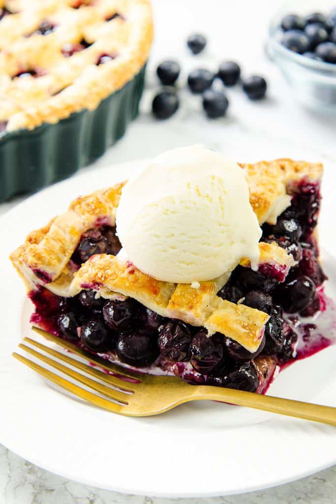A slice of homemade blueberry pie with a golden lattice crust, topped with vanilla ice cream, sits on a white plate beside a gold fork. Fresh blueberries and the rest of the blueberry pie are visible in the background.