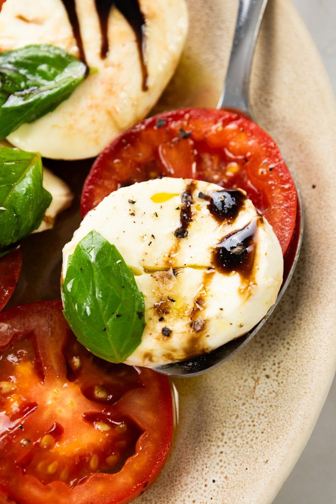 A close-up of a Classic Caprese salad with sliced tomato, fresh mozzarella, basil leaves, and a drizzle of balsamic glaze on a plate.