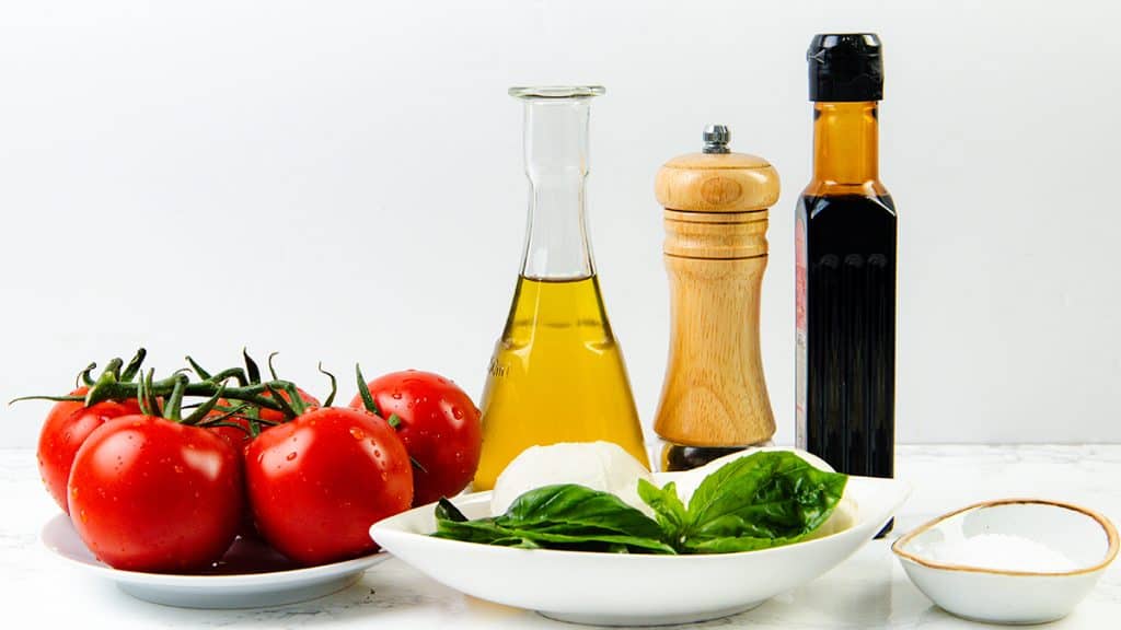 A plate of tomatoes, mozzarella, basil leaves, and a small dish of salt are artfully arranged with olive oil, balsamic vinegar, and a pepper grinder—perfect ingredients for a Classic Salad or your favorite Caprese Recipe.
