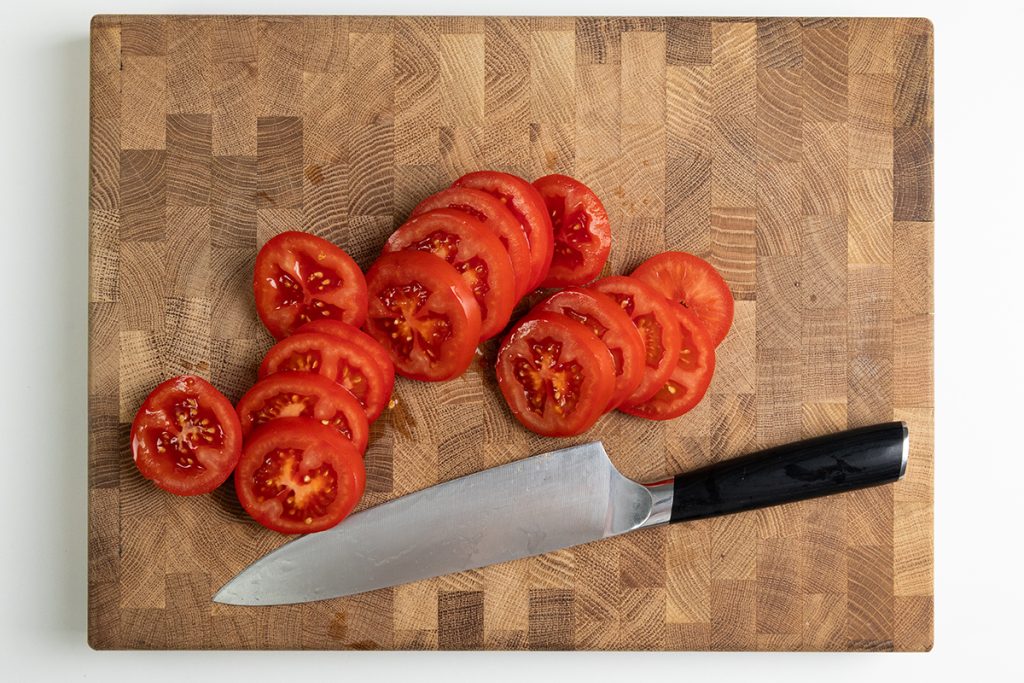 Sliced tomatoes arranged on a wooden cutting board next to a kitchen knife with a black handle, ready to be layered into a classic Caprese Salad.