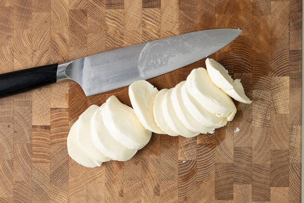 A kitchen knife with a black handle lies on a wooden cutting board next to several slices of fresh mozzarella cheese, ready to be used in a Classic Caprese Salad.