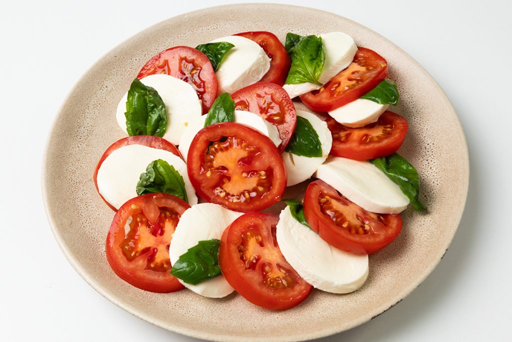 A plate of Classic Caprese Salad with sliced fresh tomatoes, mozzarella cheese, and basil leaves arranged in an overlapping pattern on a light beige plate, set against a white background.