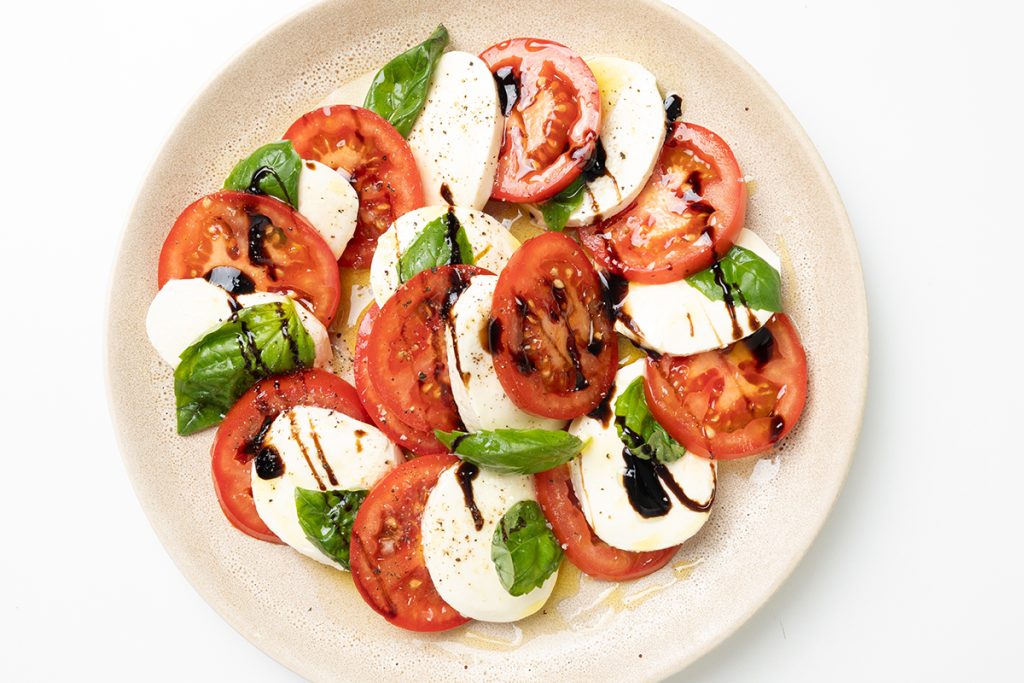 A plate of Classic Caprese Salad with sliced tomatoes, mozzarella cheese, fresh basil leaves, and a drizzle of balsamic glaze is served on a tan plate against a white background.