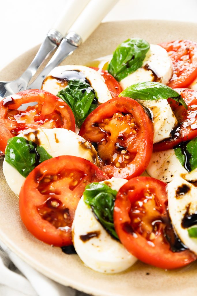 A plate of Classic Caprese Salad featuring slices of fresh tomato, mozzarella, and basil leaves, drizzled with balsamic glaze. Two forks rest on the edge—an inviting take on the timeless Caprese recipe.