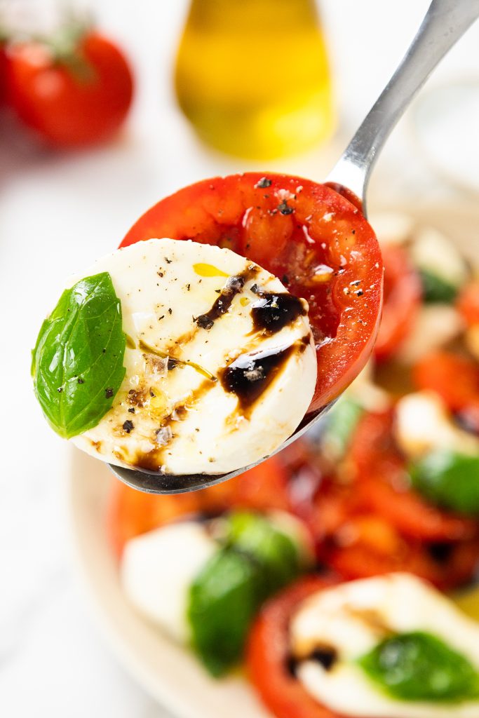 A spoon holds a slice of fresh tomato, mozzarella, and basil, drizzled with balsamic glaze—a Classic Caprese—while more Caprese Salad and an oil bottle are visible in the background.