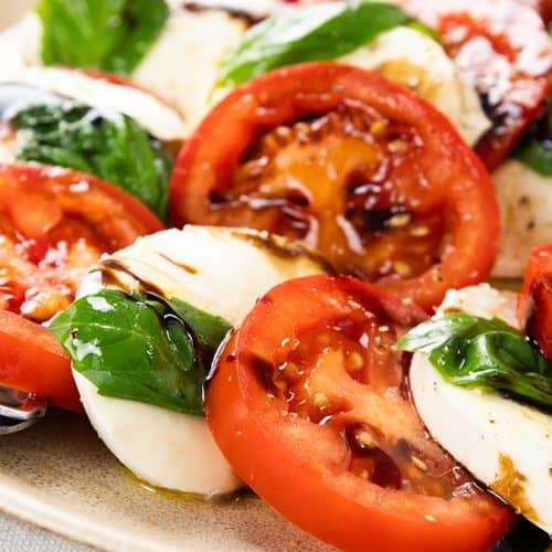 A plate of Classic Caprese Salad with fresh tomato, mozzarella cheese, and basil leaves, drizzled with balsamic glaze. Two forks rest on the edge of the plate.