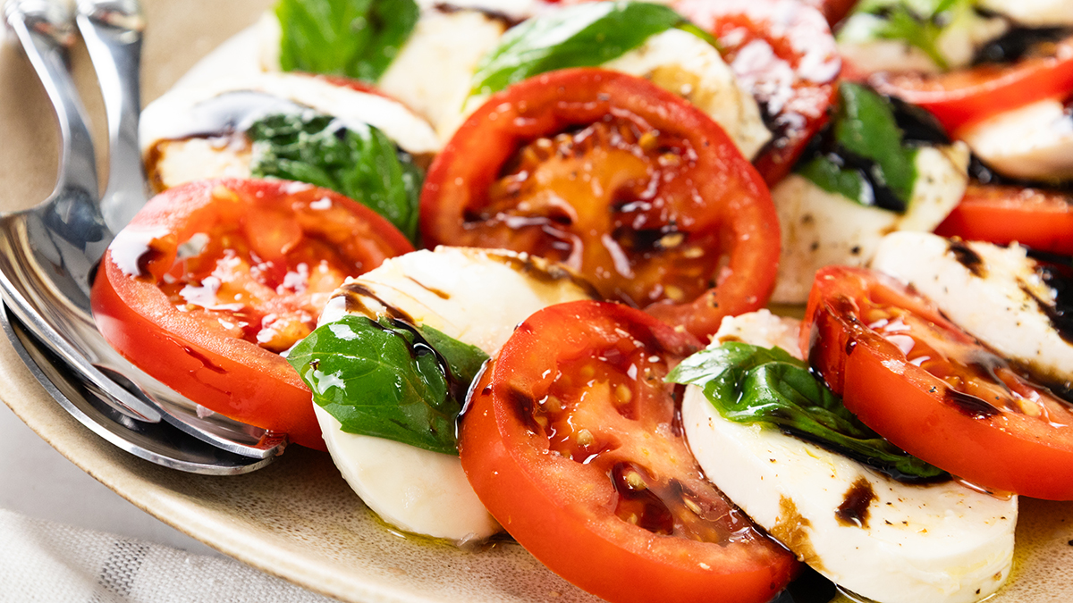 A plate of Classic Caprese Salad with fresh tomato, mozzarella cheese, and basil leaves, drizzled with balsamic glaze. Two forks rest on the edge of the plate.