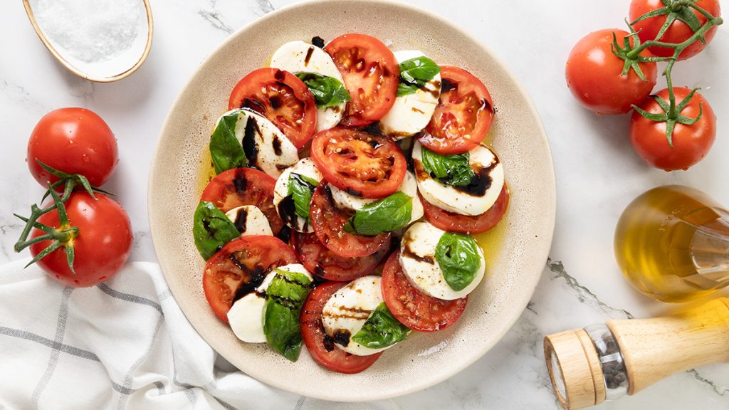 A plate of Caprese Salad featuring sliced tomatoes, fresh mozzarella, basil leaves, and balsamic glaze. Whole tomatoes, a bottle of olive oil, salt, and a pepper grinder are nearby on a marble surface.