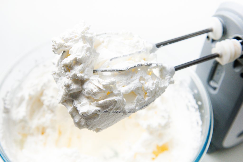 A close-up of an electric hand mixer with beaters covered in whipped cream, held above a glass bowl filled with more whipped cream&mdash;perfect for topping your Easy Lemon Pie.