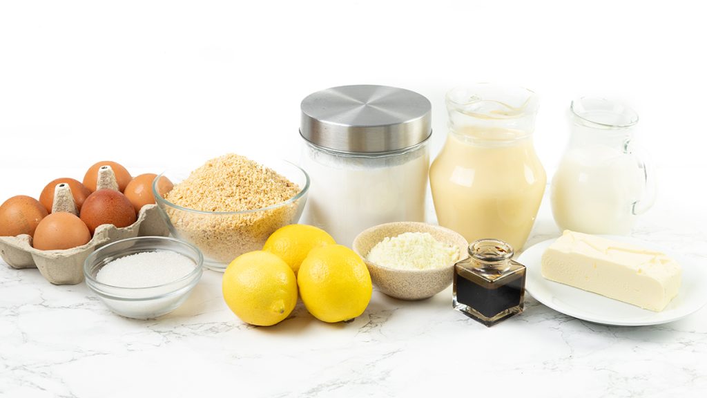 A variety of baking ingredients on a white surface, perfect for making Easy Lemon Pie, including eggs, sugar, lemons, breadcrumbs, flour, vanilla extract, butter, a jar of milk, a jar of cream, and a container of white powder.