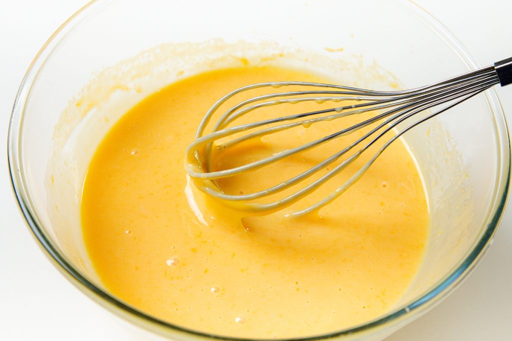A metal whisk rests in a glass bowl of smooth, yellow Easy Lemon Pie batter, set on a white background.